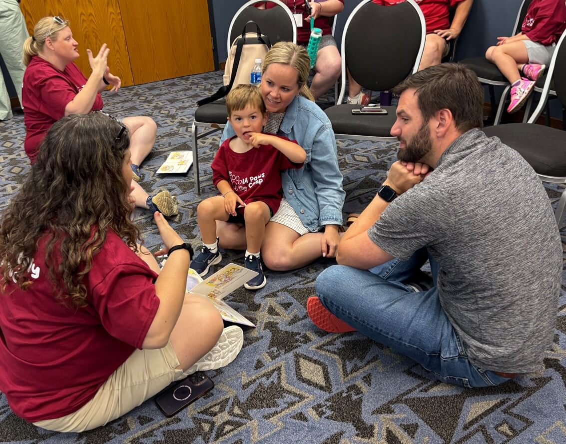 A Deaf mentor sits on the floor with two parents. One parent has a child in her lap. 